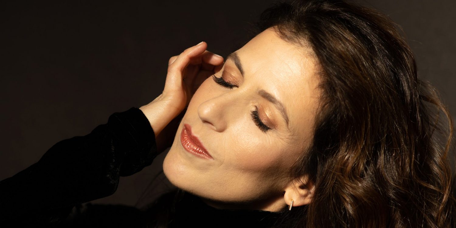 Inbal Segev wears a black velvet top and rests her hand against her face with her eyes closed and her hair flowing behind her.