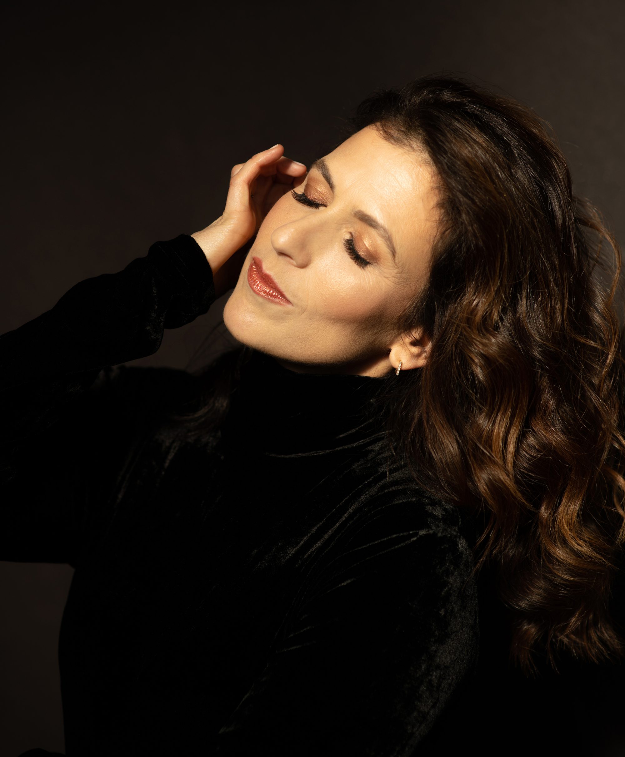Inbal Segev wears a black velvet top and rests her hand against her face with her eyes closed and her hair flowing behind her.