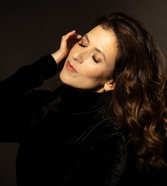 Inbal Segev wears a black velvet top and rests her hand against her face with her eyes closed and her hair flowing behind her.