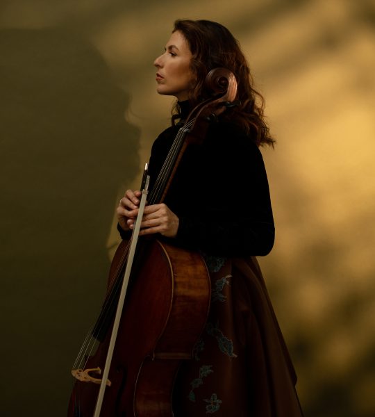 Inbal Segev stands against a light brown background, wearing a brown skirt with a blue flower pattern and a black top, while looking to the side and holding her cello.