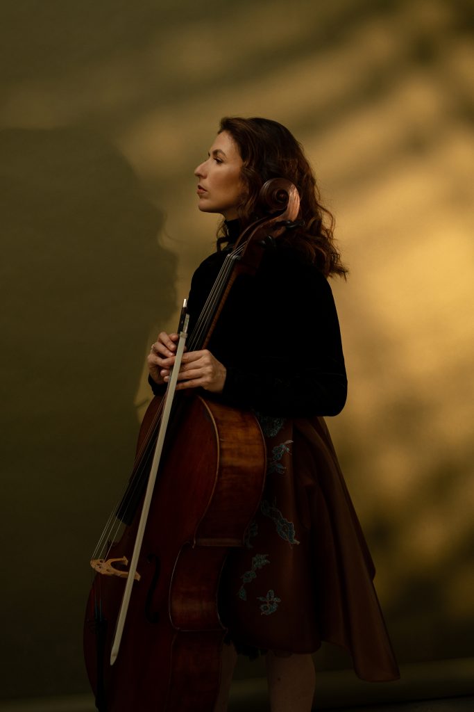Inbal Segev stands against a light brown background, wearing a brown skirt with a blue flower pattern and a black top, while looking to the side and holding her cello.