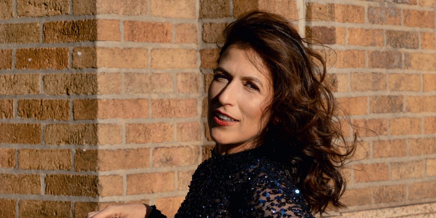 Inbal Segev stands in front of a brick wall wearing a blue sequined gown, with her cello case leaning against her.