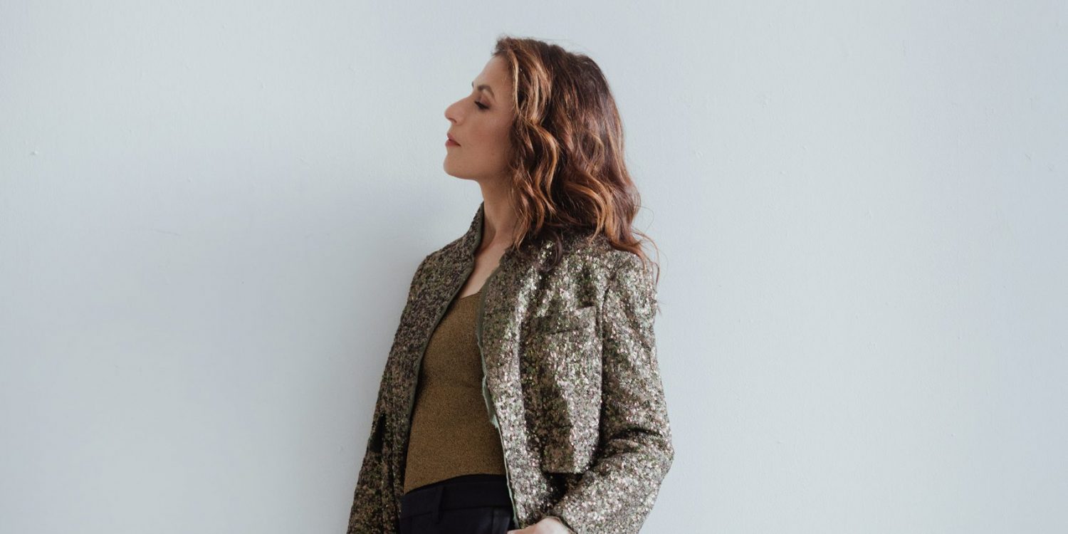 Inbal Segev leans against a while wall wearing dark pants, and a brown patterned blazer.