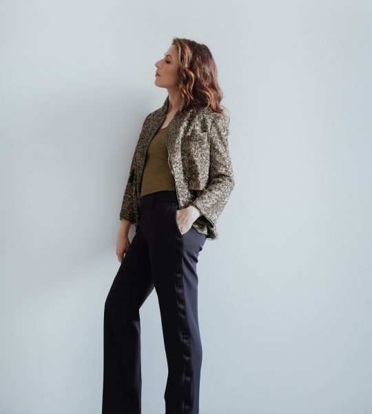 Inbal Segev leans against a while wall wearing dark pants, and a brown patterned blazer.