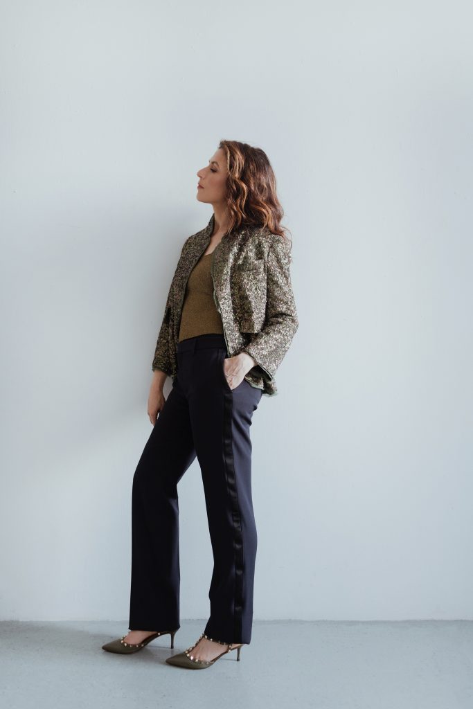 Inbal Segev leans against a while wall wearing dark pants, and a brown patterned blazer.