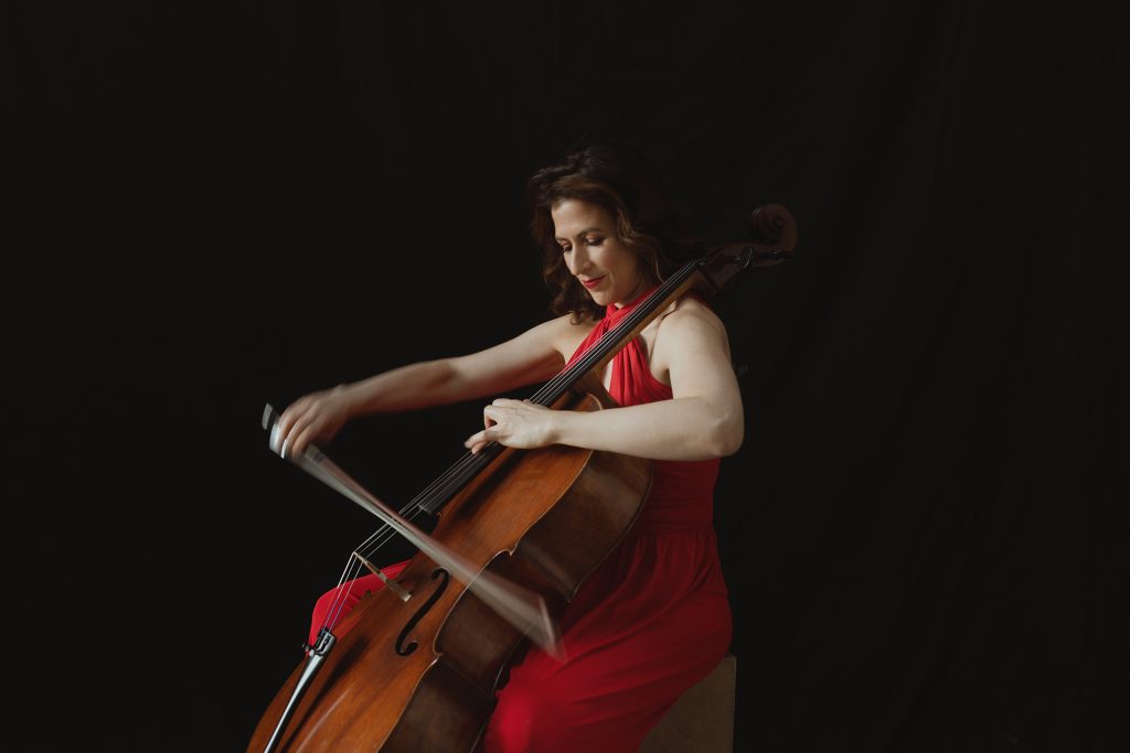 Inbal Segev, in a flowing red gown, sits on a chair and plays her cello.