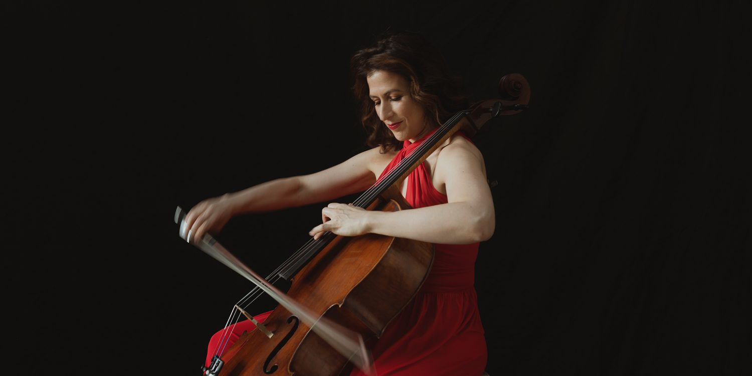 Inbal Segev, in a flowing red gown, sits on a chair and plays her cello.