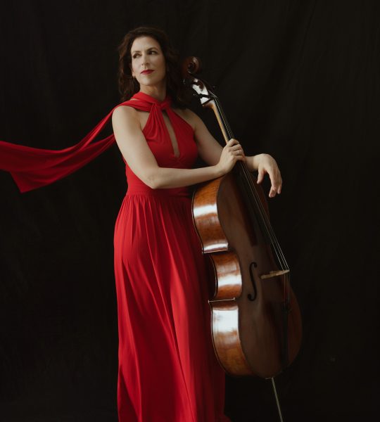 Inbal Segev stands against a dark backdrop wearing a flowing red gown and resting her cello against her side.