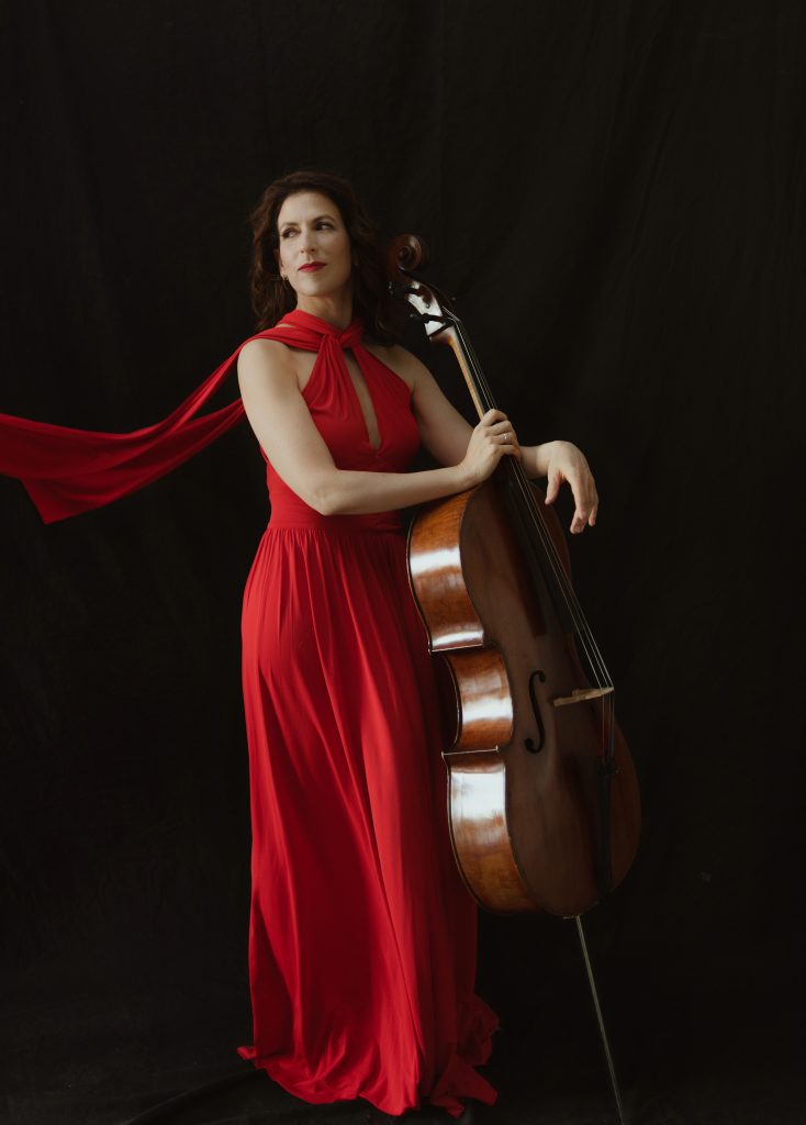 Inbal Segev stands against a dark backdrop wearing a flowing red gown and resting her cello against her side.