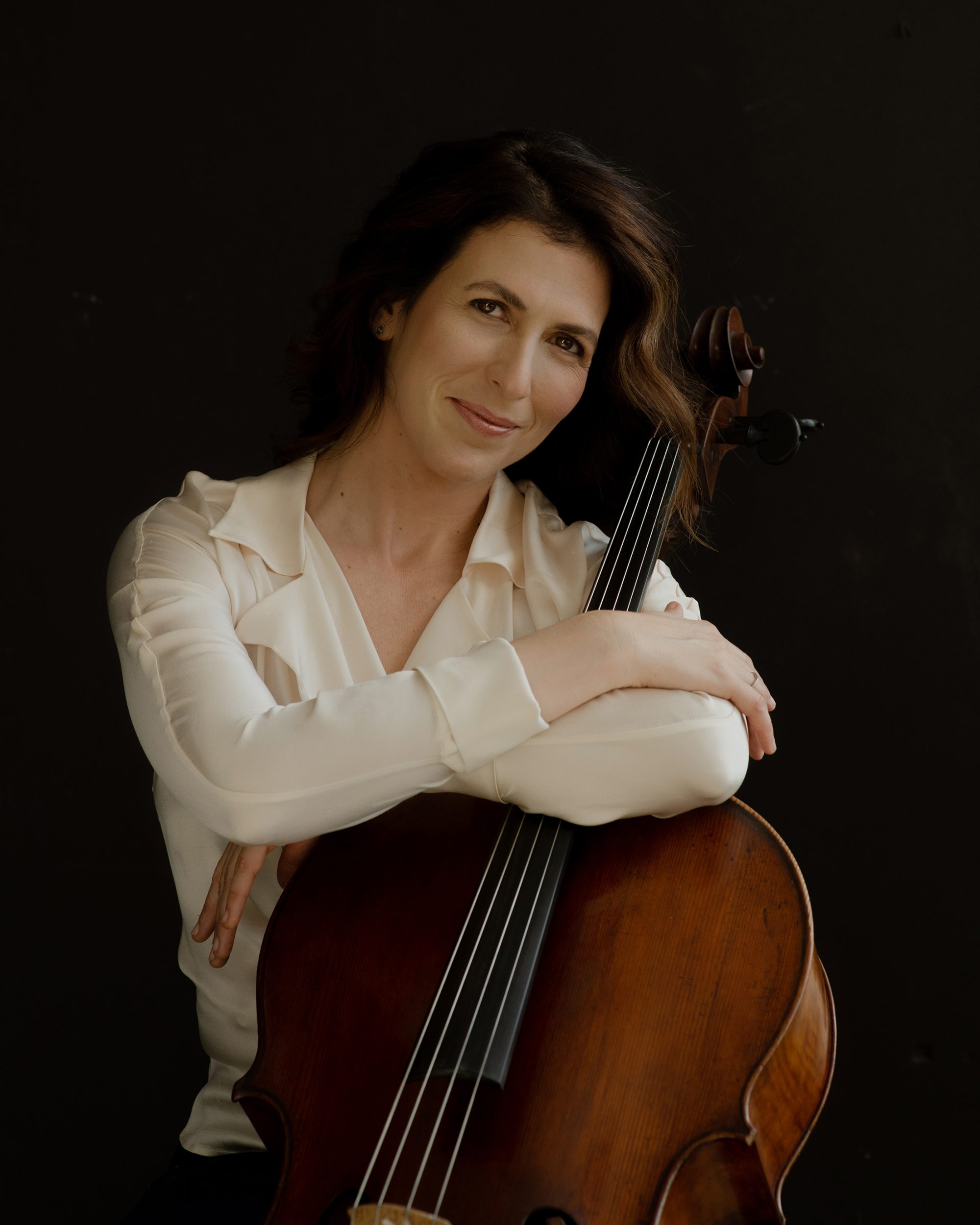 Inbal Segev hugs her cello and smiles, wearing a white blouse against a black background.