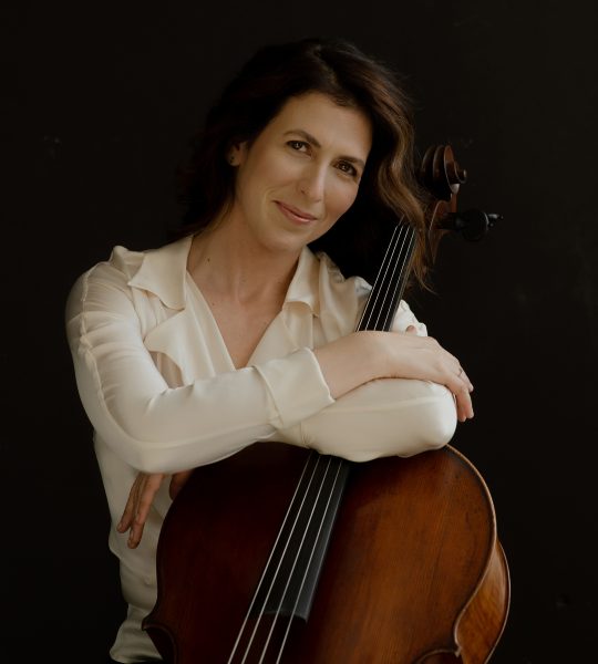 Inbal Segev hugs her cello and smiles, wearing a white blouse against a black background.