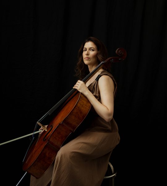 Inbal Segev sits on a stool holding her cello and bow, wearing a sleeveless loose brown dress and looking serious.