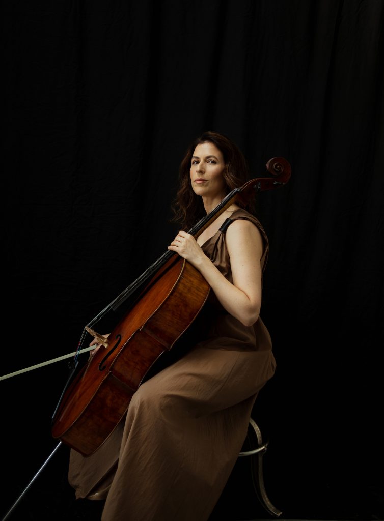 Inbal Segev sits on a stool holding her cello and bow, wearing a sleeveless loose brown dress and looking serious.