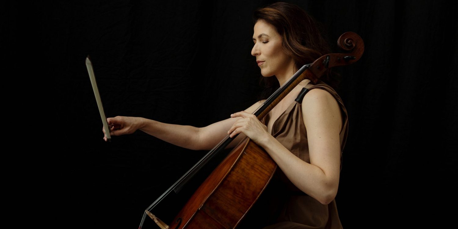 Inbal Segev sits on a stool, playing her cello in a loose, sleeveless brown dress against a black background.