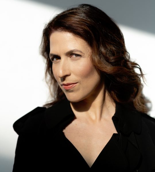 Inbal Segev wears a black jacket against a white background, looking serious with a slight smile.