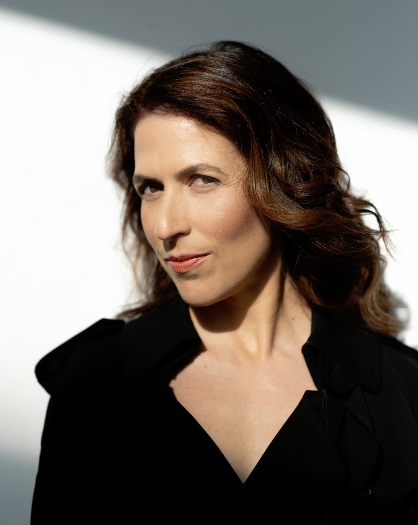 Inbal Segev wears a black jacket against a white background, looking serious with a slight smile.