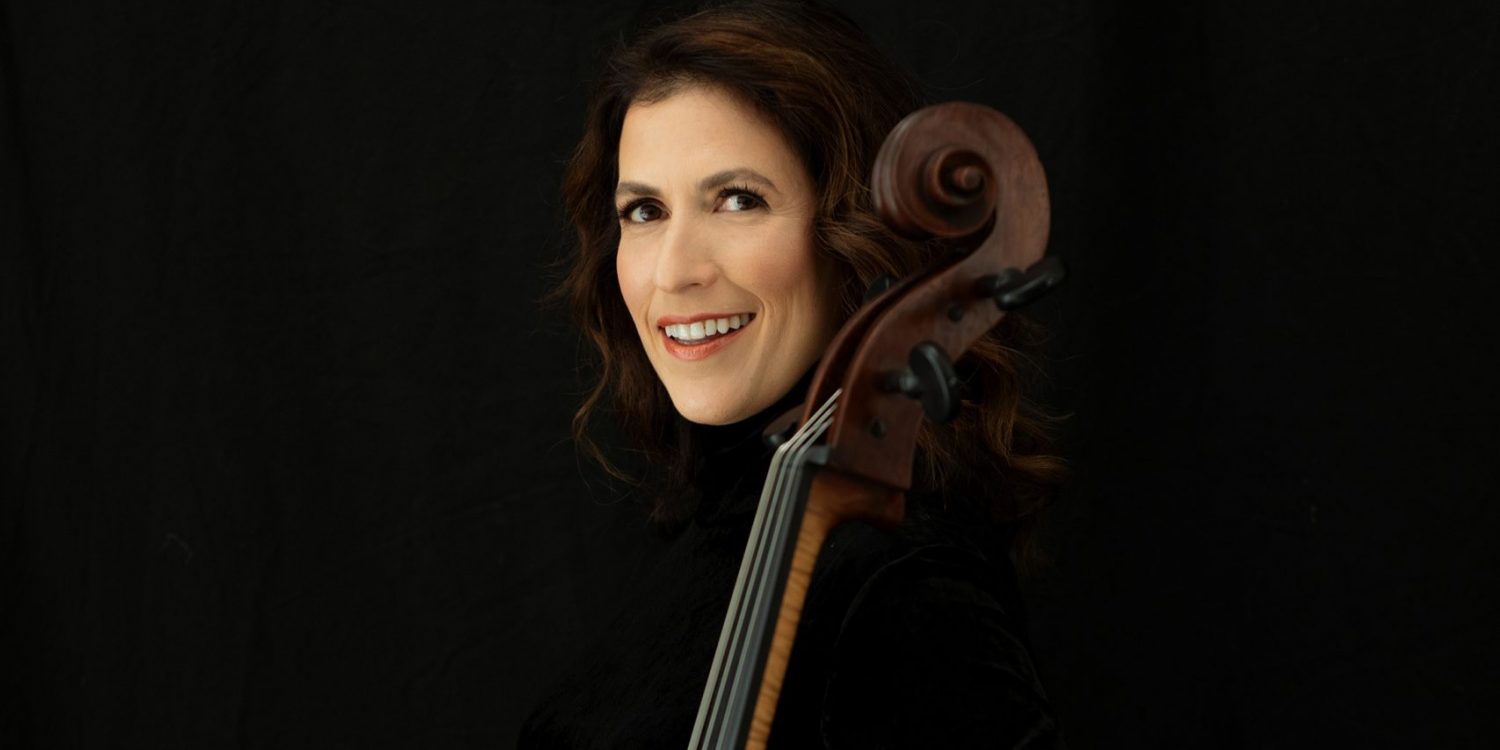 Inbal Segev stands with her cello against a black background smiling and wearing a black top and a brown skirt with a blue flower pattern.