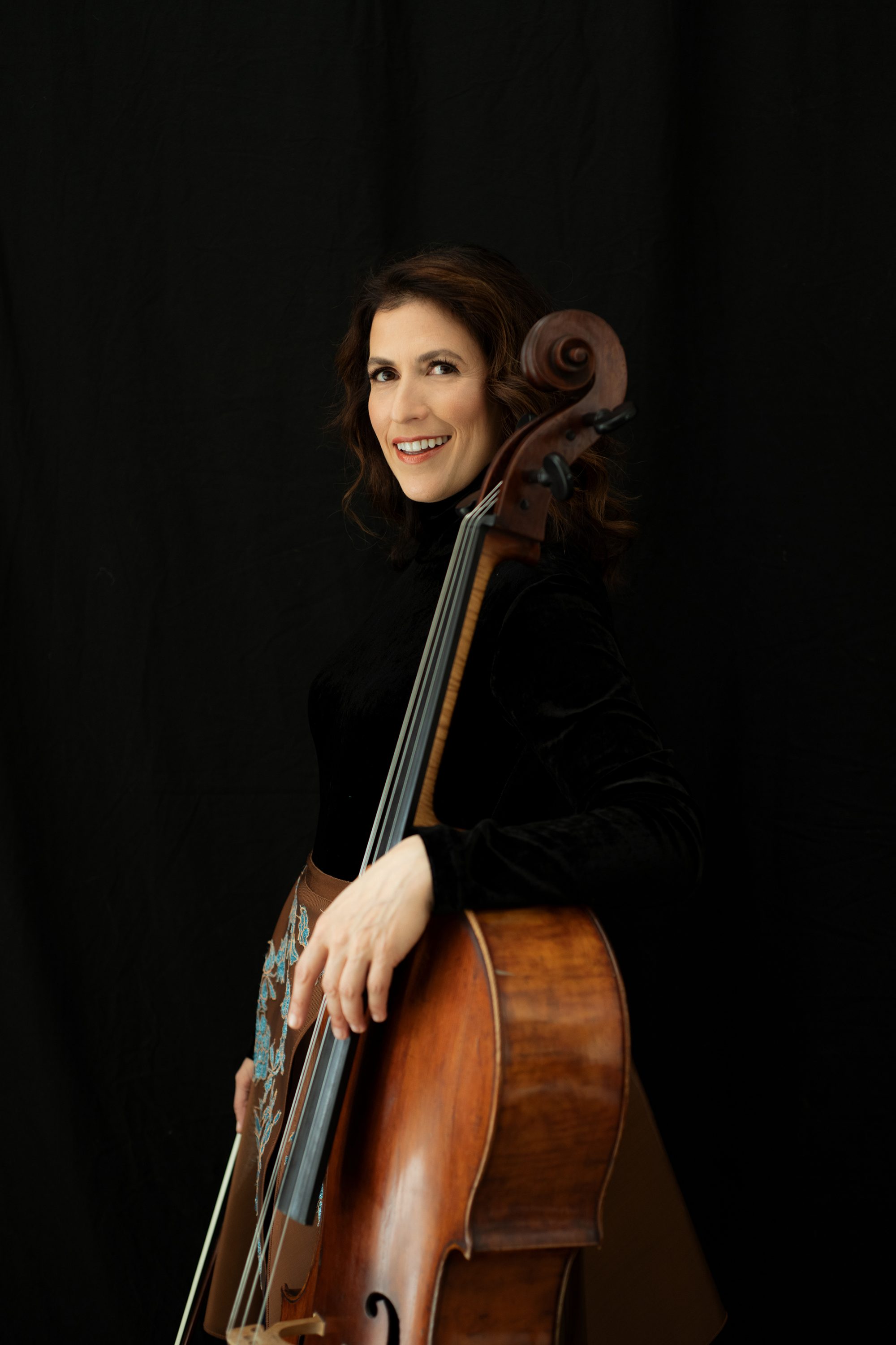 Inbal Segev stands with her cello against a black background smiling and wearing a black top and a brown skirt with a blue flower pattern.